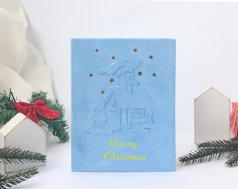 May include: A light blue Christmas card with an embossed house and star design. The words "Merry Christmas" are in yellow. Decorative wooden houses, a red bow, and pine branches are in the background.