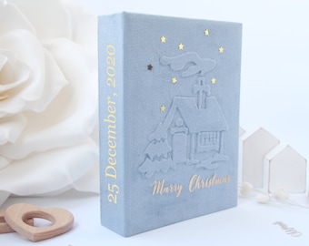 May include: A light blue velvet-textured book with gold foil text on the spine reading 25 December, 2020. The front cover features an embossed house with smoke, small gold star accents, and the gold foil text Marry Christmas at the bottom.