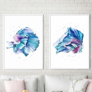 Könnte beinhalten: Zwei Aquarellbilder von blauen Betta-Fischen mit rosa Akzenten. Die Fische sind in einer dynamischen Pose mit ausgebreiteten Flossen dargestellt. Die Bilder sind in weißen Rahmen gerahmt und an einer weißen Backsteinwand aufgehängt.