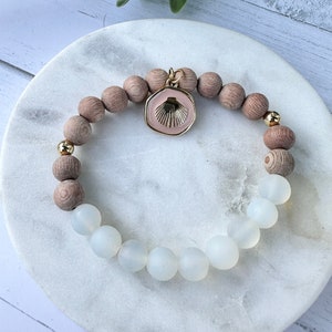 Könnte beinhalten: Ein Armband aus Holz- und weißen Perlen mit einem goldenen Anhänger, der eine rosa Muschel zeigt. Das Armband liegt auf einem weißen Teller.