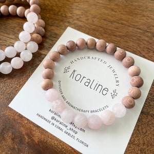 Op de afbeelding: Een houten kralen armband met roze rozenkwarts kralen. De armband is handgemaakt en heeft de tekst "HANDCRAFTED JEWELRY Koraline DIFFUSER AND AROMATHERAPY BRACELETS koraline...etsy.com @koraline_shop HANDMADE IN CORAL GABLES, FLORIDA".