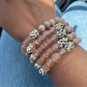 May include: A stack of bracelets with pink and white beads. The beads are made of glass and wood. The bracelets are stacked on a person's wrist.