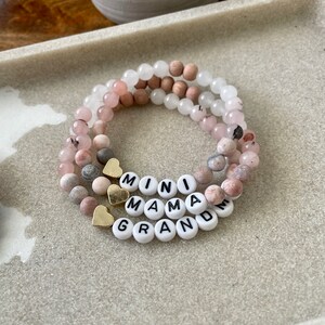 May include: Three layered bracelets with pink, white and brown beads. The bracelets spell out "Mini Mama Grandma" with gold heart charms.