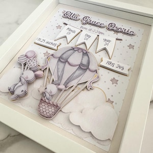 Op de afbeelding: Een ingelijste geboorteaankondiging met een luchtballonthema. Het kunstwerk bevat de naam van de baby, de geboortedatum, tijd, lengte (48.9 cm) en gewicht (3.2 kg). Het ontwerp toont een luchtballon, wolken en sterren in zachte pastelkleuren.