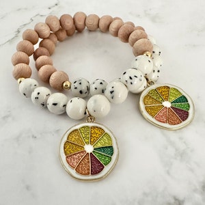 Op de afbeelding: Twee armbanden met houten en wit gespikkelde kralen. Elke armband heeft een bedel in de vorm van een kleurrijke citrusschijf.