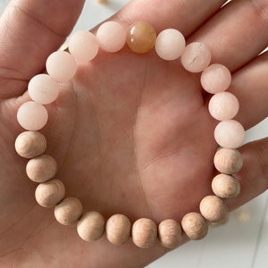 May include: A bracelet made of light pink and natural wood beads. The bracelet has a light orange bead in the centre.