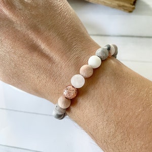Pode incluir: Um bracelete feito de pedras naturais em tons de cinza, branco e rosa. O bracelete apresenta uma conta de ouro rosa com uma superfície texturizada.