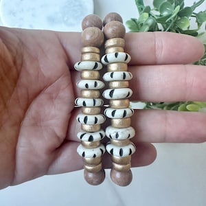 Peut inclure: Deux bracelets en perles de bois avec des accents dorés et blancs. Les perles sont ornées de rayures noires et blanches.