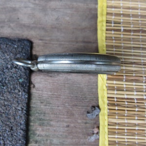 Puede incluir: Un reloj de bolsillo plateado con un bisel texturizado y acanalado y un peque&ntilde;o lazo para una cadena. El reloj est&aacute; sobre una superficie de madera, con una secci&oacute;n de asfalto gris oscuro y una estera de bamb&uacute; en el fondo.