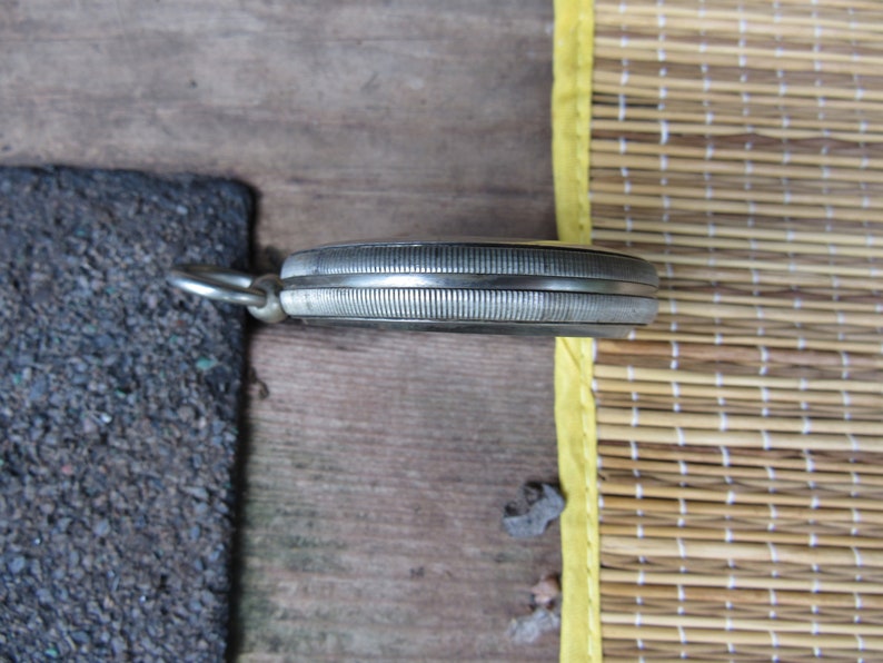 Puede incluir: Un reloj de bolsillo plateado con un bisel texturizado y acanalado y un peque&ntilde;o lazo para una cadena. El reloj est&aacute; sobre una superficie de madera, con una secci&oacute;n de asfalto gris oscuro y una estera de bamb&uacute; en el fondo.