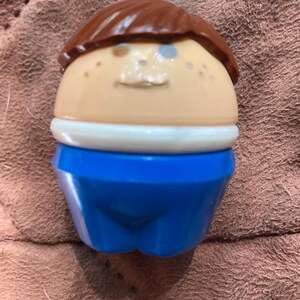 May include: A small, egg-shaped plastic toy figure with a smooth, tan face, brown molded hair, and a white collar. The figure wears bright blue pants and features simple, painted facial details including eyes and a small mouth on its rounded head.