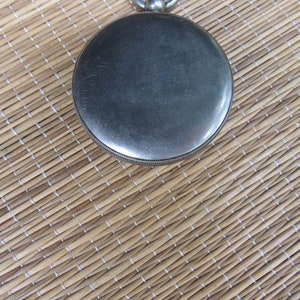 Puede incluir: Primer plano de un reloj de bolsillo plateado con esfera circular y borde estriado. El reloj se encuentra sobre un fondo tejido marr&oacute;n claro, creando un contraste de texturas y colores. Un reloj cl&aacute;sico.