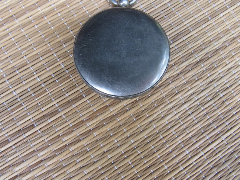 Puede incluir: Primer plano de un reloj de bolsillo plateado con esfera circular y borde estriado. El reloj se encuentra sobre un fondo tejido marr&oacute;n claro, creando un contraste de texturas y colores. Un reloj cl&aacute;sico.