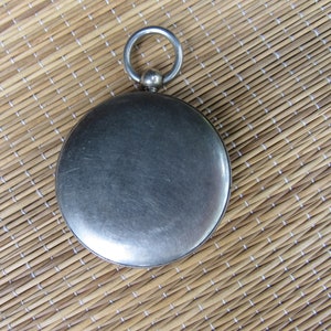 Puede incluir: Un reloj de bolsillo redondo, de color plateado, con un lazo en la parte superior. El reloj tiene una superficie lisa y reflectante y un borde decorativo. El fondo es una estera tejida de color marr&oacute;n claro.