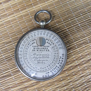 Puede incluir: Un fot&oacute;metro infalible Wynne's vintage, un dispositivo circular con una carcasa de metal y una cubierta de vidrio. La esfera presenta n&uacute;meros y texto, incluyendo "Actino Time" y "Exposure in Seconds or Minutes". Tiene un anillo para colgar.