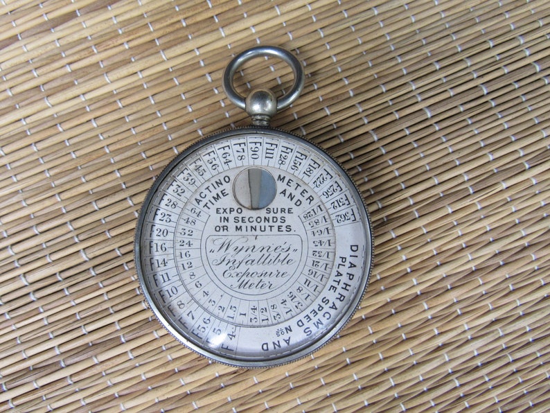 Puede incluir: Un fot&oacute;metro infalible Wynne's vintage, un dispositivo circular con una carcasa de metal y una cubierta de vidrio. La esfera presenta n&uacute;meros y texto, incluyendo "Actino Time" y "Exposure in Seconds or Minutes". Tiene un anillo para colgar.