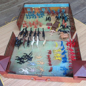 May include: A vintage toy playset featuring a Wild West theme. The set includes miniature plastic figures of cowboys, Native Americans, horses, and buildings. The figures are in various colors, including blue, red, black, and tan. The playset is housed in a brown case.