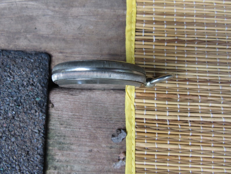 Puede incluir: Un reloj de bolsillo plateado con un bisel acanalado y un lazo para una cadena. El reloj se encuentra sobre un fondo de superficie de madera y una estera de bamb&uacute; tejida con ribete amarillo.