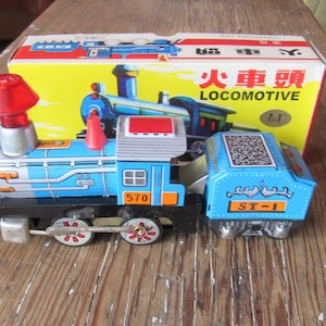 May include: A blue and silver toy train with a red smokestack and a black coal car. The train has a number "570" on the side and the words "ST-1" on the coal car. The train is in its original box, which is yellow with red and black lettering. The box says "Locomotive" in English and Chinese.