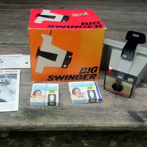 May include: Vintage Big Swinger camera set with box, flashbulbs, and manuals. The box is orange and red with the words "BIG SWINGER" printed on it. The camera is gray and black.