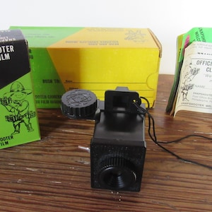 May include: A black Snapshooter camera with a black film cartridge box, a yellow and green cardboard box, and a paper membership card. The film box features a cartoon character and text reading Snapshooter Club Film 126 Cartridge Black & White.