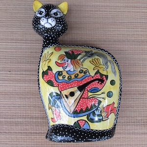 May include: A black and white ceramic cat figurine with a yellow clown playing a musical instrument on its side. The clown is wearing a red and white polka dot costume and a yellow hat.