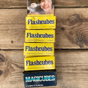 May include: A stack of vintage Flashcubes and Magicubes boxes. The Flashcubes boxes are yellow with blue text, advertising 12 guaranteed flashes and 3 cubes. The Magicubes box is blue with white and turquoise text, for 3 cubes and 12 flashes.