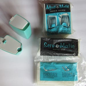May include: Vintage kitchenware set featuring a turquoise napkin holder and butter dish, alongside packaged items labeled "Adjust-O-Matic Napkin Holder" and "Serv-O-Matic Butter Dish." The packaging has a retro design with teal and black text.
