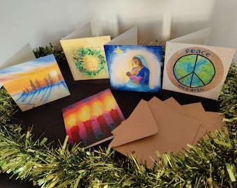 Puede incluir: Un conjunto de cinco tarjetas de felicitación cuadradas con varias ilustraciones festivas, incluyendo un bosque al atardecer, una corona verde, una escena de natividad, cuatro velas encendidas y un globo terráqueo con el texto Peace on Earth, junto a sobres marrones.