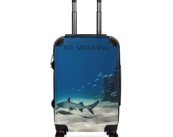 May include: A rolling suitcase featuring an underwater scene with a shark swimming near the ocean floor. The suitcase has a telescoping handle and wheels. The words "NO SWIMMING" are printed at the top of the suitcase.