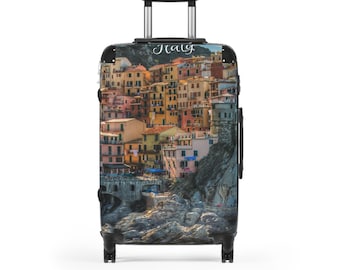Puede incluir: Una maleta con ruedas con una imagen vibrante de un pueblo costero italiano. La maleta tiene un asa y un marco negros, con la palabra "Italy" en escritura en la parte superior. Los edificios están pintados en tonos de amarillo, rosa y naranja.
