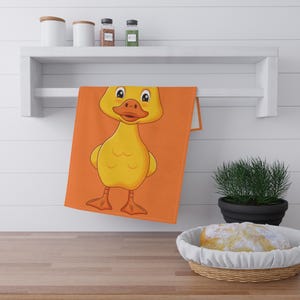 Op de afbeelding: Oranje keukentowel met een cartoon eend ontwerp. De eend is geel met een oranje snavel en poten. De handdoek hangt aan een witte plank. De achtergrond is fel oranje.