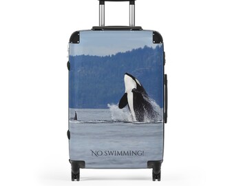 May include: A hard-shell rolling suitcase featuring a graphic print of an orca breaching the water surface with a distant forest background. The text NO SWIMMING! is printed in black at the bottom. The luggage has a black handle and wheels.