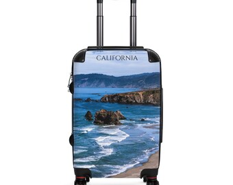May include: A rolling suitcase featuring a scenic coastal image of California. The image shows a blue ocean, rocky cliffs, and a sandy beach under a clear sky. The word "CALIFORNIA" is printed at the top of the suitcase.