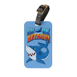 Puede incluir: Etiqueta de equipaje azul con un tiburón de dibujos animados y el texto "SHARK ATTACK!" en letras naranja y amarillas.