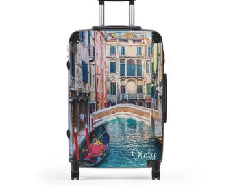Puede incluir: Una maleta con ruedas con una imagen colorida de Venecia, Italia, con una góndola en el agua y edificios a lo largo del canal. La palabra "Italy" está escrita en cursiva. La maleta tiene un asa y ruedas negras.