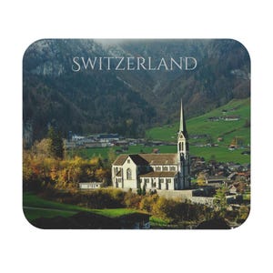 May include: A rectangular coaster featuring a scenic view of Switzerland. The image shows a church with a tall steeple in a valley, surrounded by green hills and a village. The word "SWITZERLAND" is printed at the top.