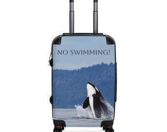 May include: A rolling suitcase featuring a graphic of a breaching orca whale. The suitcase has a black handle and wheels. The words "NO SWIMMING!" are printed across the top of the suitcase.