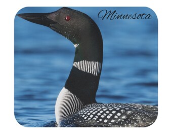May include: A black and white loon with red eyes swimming in blue water. The text "Minnesota" is written in black cursive at the top right of the image.