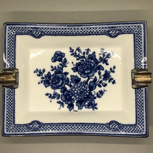 Könnte beinhalten: Ein weißer Aschenbecher aus Porzellan mit einem blauen und weißen Blumendesign und goldenen Akzenten. Der Aschenbecher hat eine quadratische Form mit abgerundeten Ecken und einem erhöhten Rand.