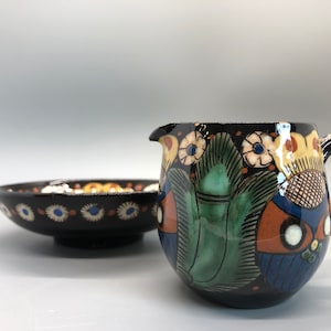 Könnte beinhalten: Ein schwarzes Keramik-Krug- und Schüssel-Set mit farbenfrohen floralen und geometrischen Designs. Der Krug hat einen Griff und einen Ausguss, und die Schüssel ist flach. Das Set hat blaue, grüne, orange und weiße Akzente.