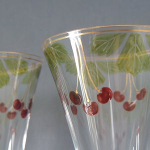 Könnte beinhalten: Zwei klare Champagnerflöten aus Glas mit goldener Verzierung und bemalten Kirschmotiven. Die Kirschen sind rot und die Blätter sind grün.
