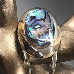 Peut inclure: Un bracelet en argent avec une grande incrustation ovale en nacre d'ormeau. La nacre présente des couleurs irisées bleues, vertes et violettes.