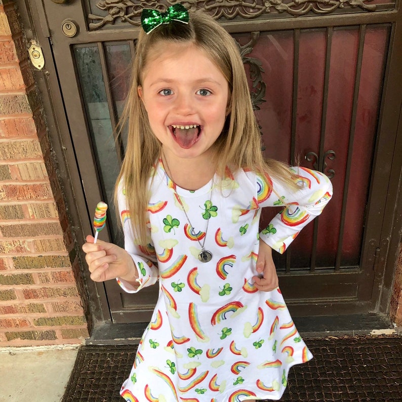 May include: A young girl wearing a white dress with a rainbow and shamrock pattern. She is holding a colorful lollipop and has a green bow in her hair.