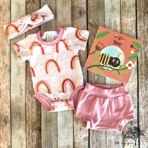May include: A baby outfit set with a pink headband, a white bodysuit with rainbow and heart print, pink bloomers, and a board book titled "Talking Friends Nature: Bee".