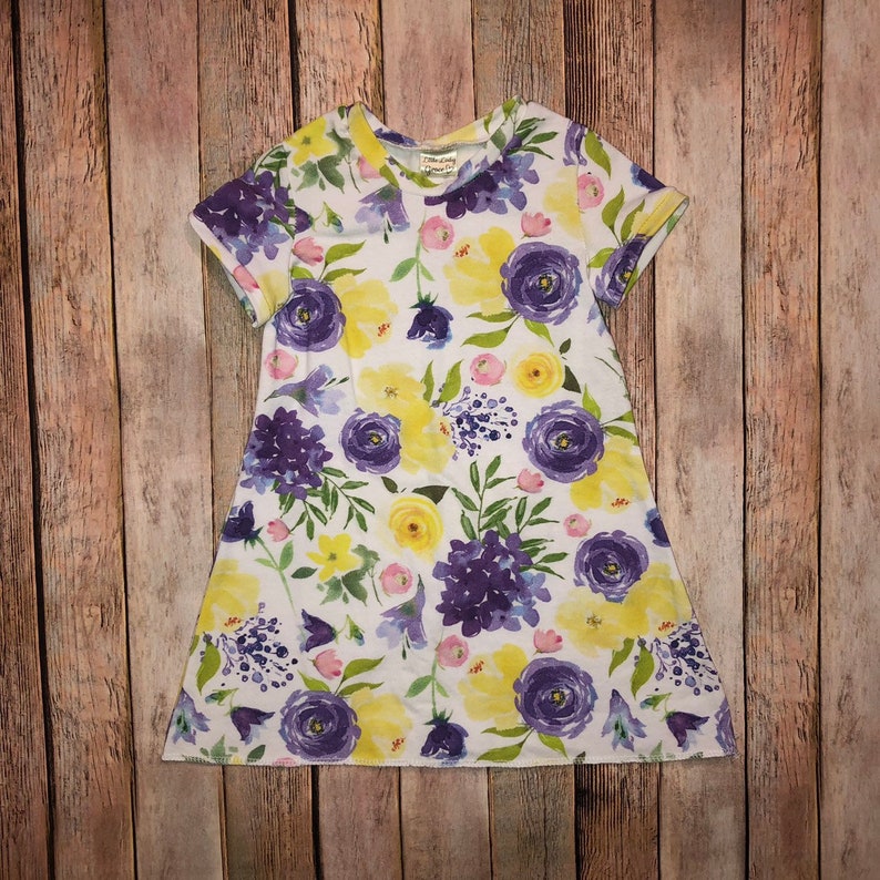 May include: A white dress with a floral print featuring yellow and purple flowers. The dress has short sleeves and a round neckline.