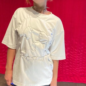 Pode incluir: Camiseta branca com um desenho de caveira costurada. A camiseta de manga curta tem um corte folgado. A modelo usa calças listradas em preto e branco e um colar gargantilha prateado com pontas. O fundo é vermelho liso.