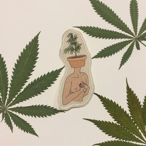 Può includere: Una toppa ricamata raffigurante una figura nuda con una testa a forma di vaso, con una pianta di cannabis che ne cresce. La toppa è circondata da foglie verdi di cannabis su sfondo bianco.