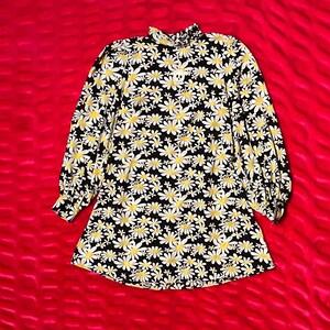 May include: A long-sleeved dress with a black background and a daisy floral pattern in white and yellow. The dress has a high neckline and a slightly flared silhouette. The fabric appears to be a lightweight material.