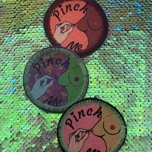 May include: Three circular embroidered patches featuring a graphic illustration of a hand pinching a nipple. Each patch displays the text "Pinch Me" in black script. The designs vary in color, featuring orange, purple, green, and pink tones.